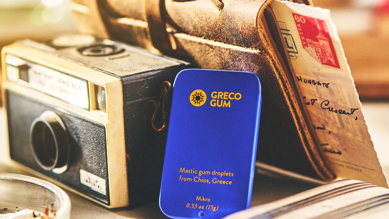 A tin of Greco Gum sits next to a vintage camera and leather journal