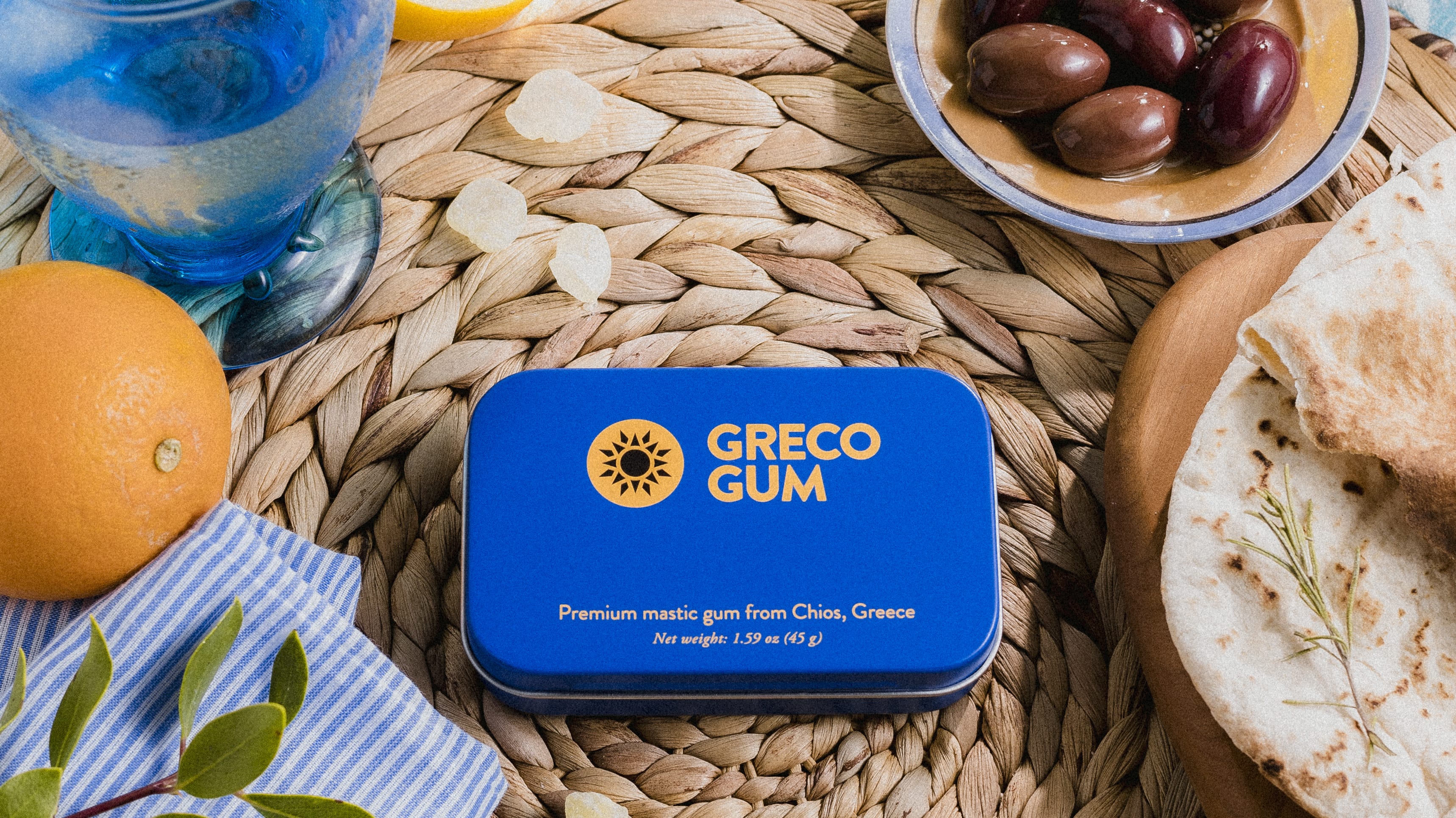 A blue tin of Greco Gum sits in the middle of a woven placemat next to a fresh Greek meal.