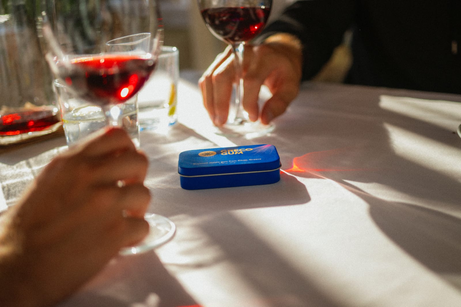 A tin of Greco Gum between glasses of wine at a white tablecloth dinner
