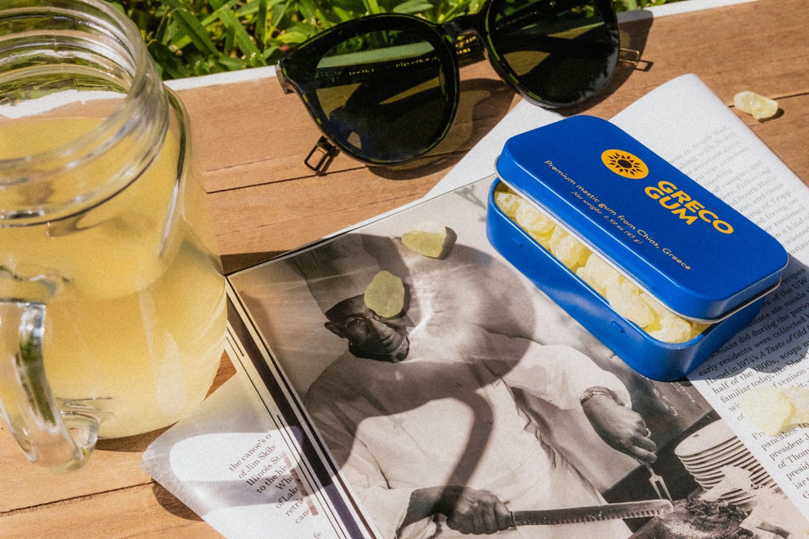 A blue tin of Greco Gum sits on top of a black and white magazine next to a mason jar and sunglasses