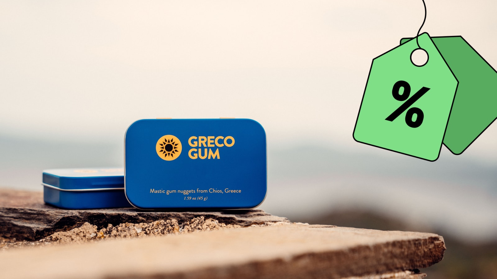 Two blue Greco Gum tins on the ledge of a stone wall with a discount tag hanging from the right side of the image.