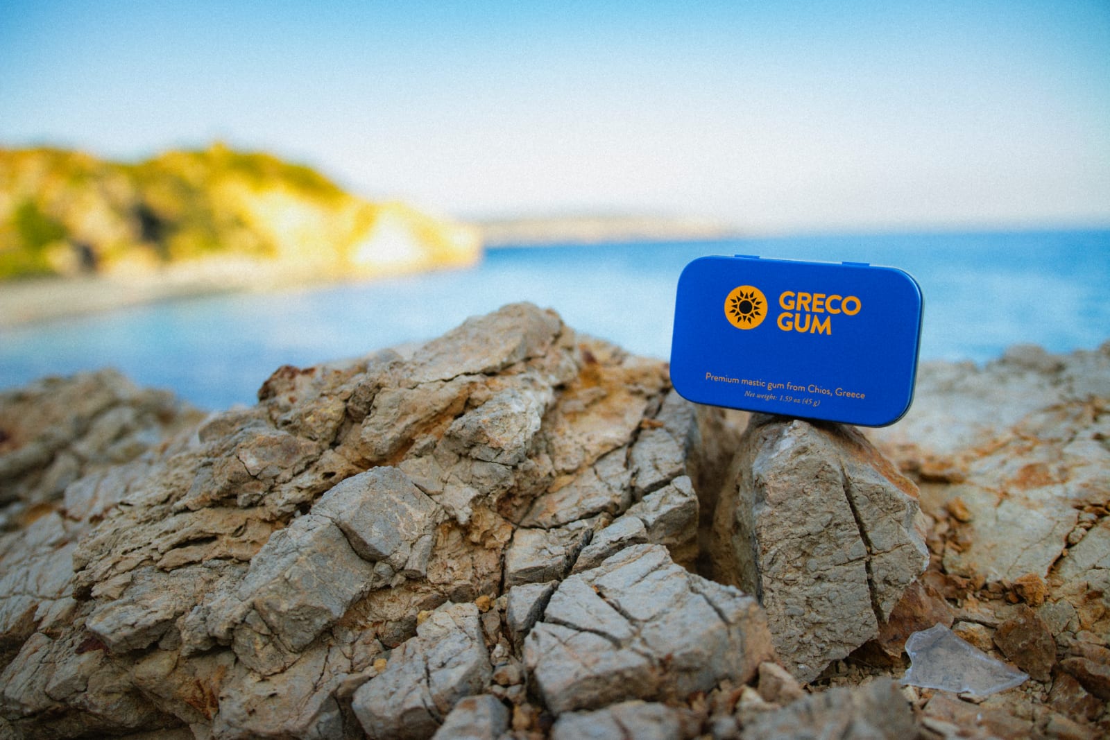 A blue tin of Greco Gum mastic gum nuggets perched atop some cliffs by the ocean in Chios, Greece.