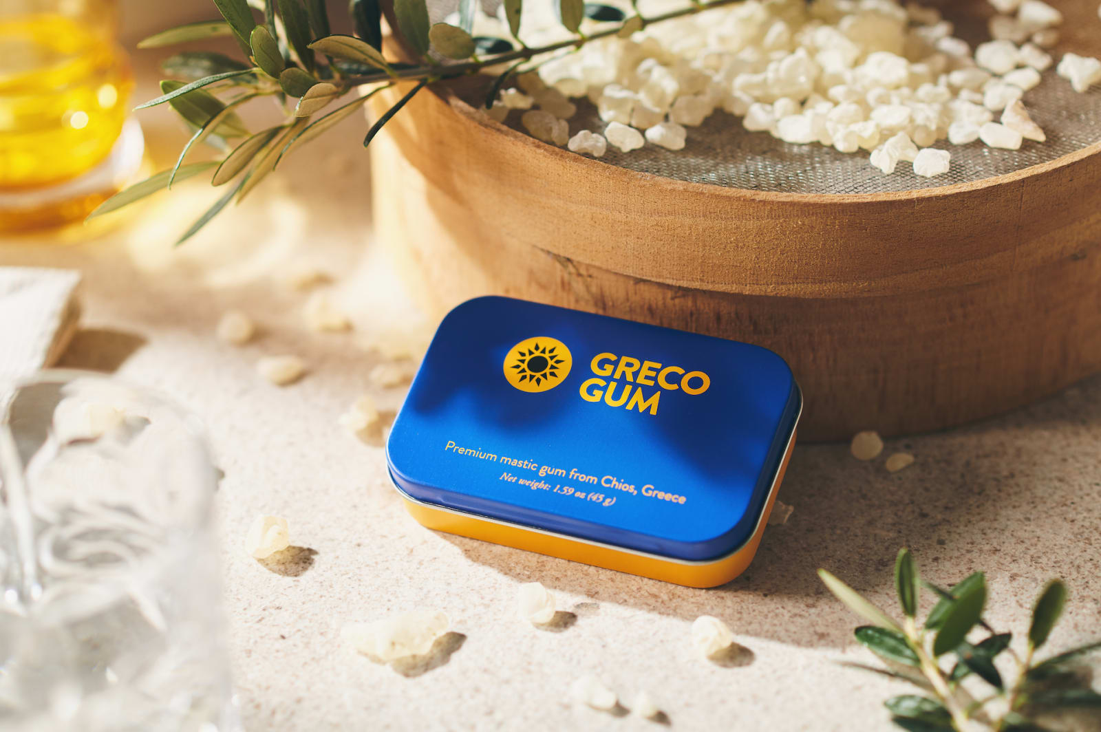 A tin of Greco Gum rests on a table with nuggets of gum sprinkled around it and more nuggets in the background.