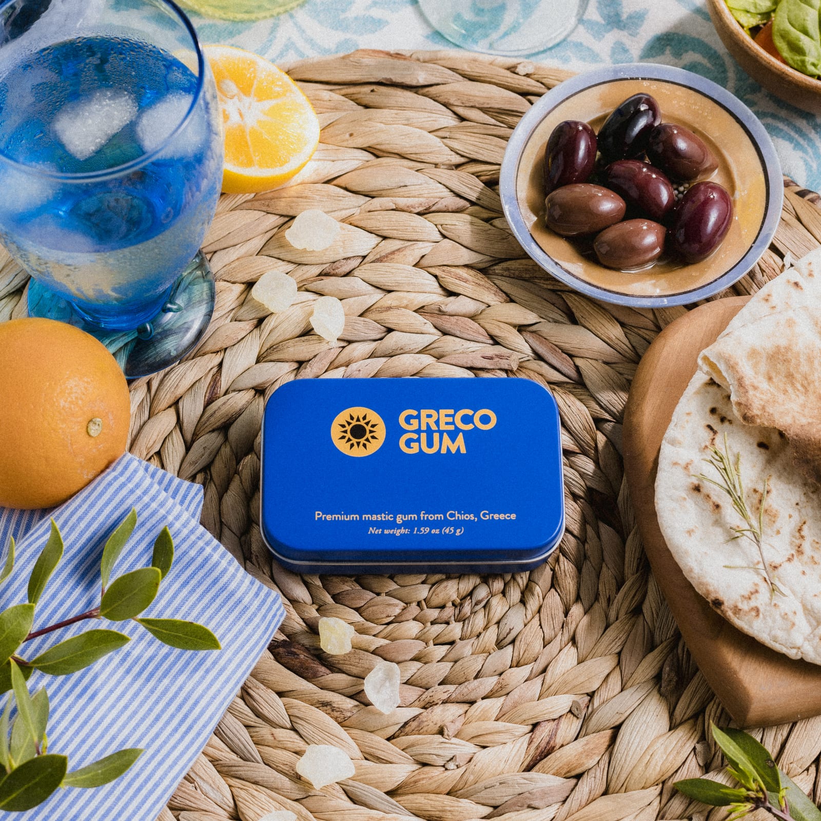 A blue tin of Greco Gum sits in the middle of a woven placemat next to a fresh Greek meal.