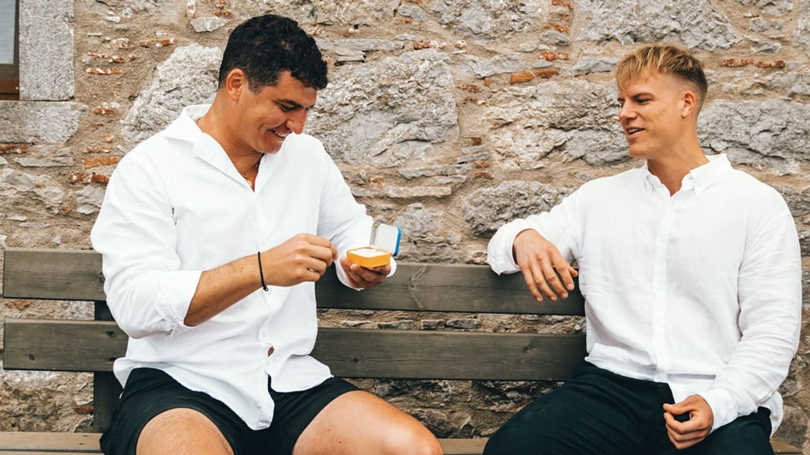 Two men in white shirts sitting on a bench laughing while picking out a piece of Greco Gum from a blue tin.