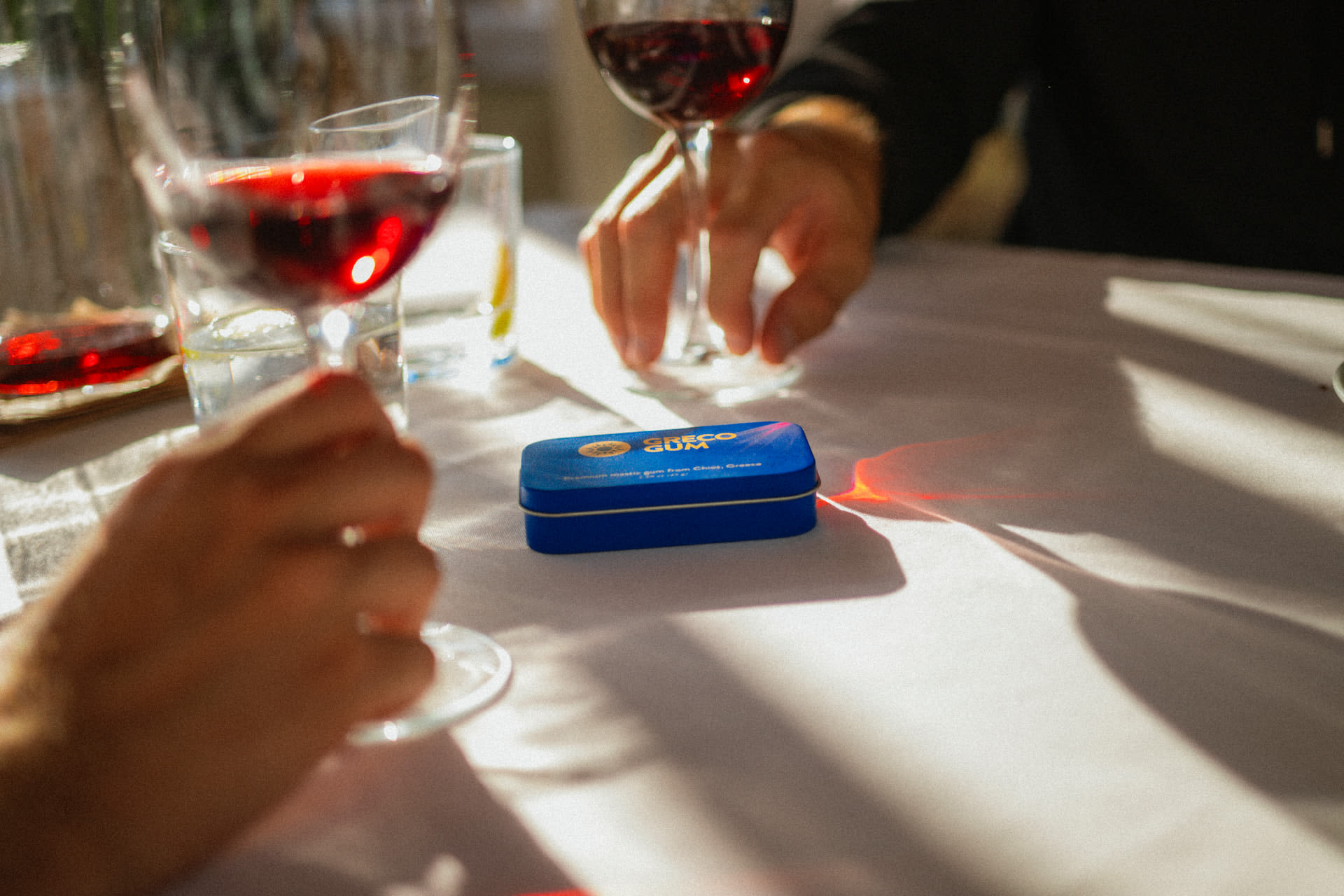 A tin of Greco Gum between glasses of wine at a white tablecloth dinner