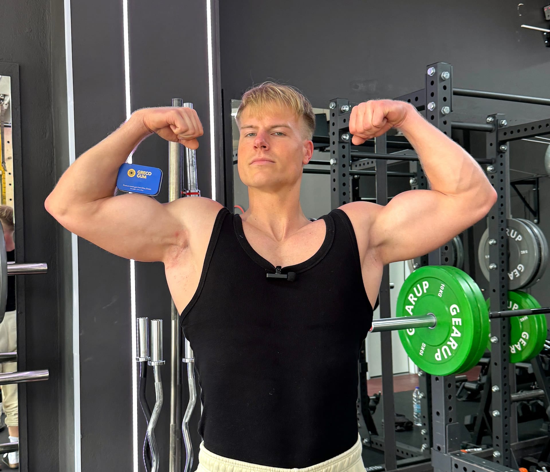 A muscular man flexes his biceps in the gym, with a tine of Greco Gum between his bicep and forearm