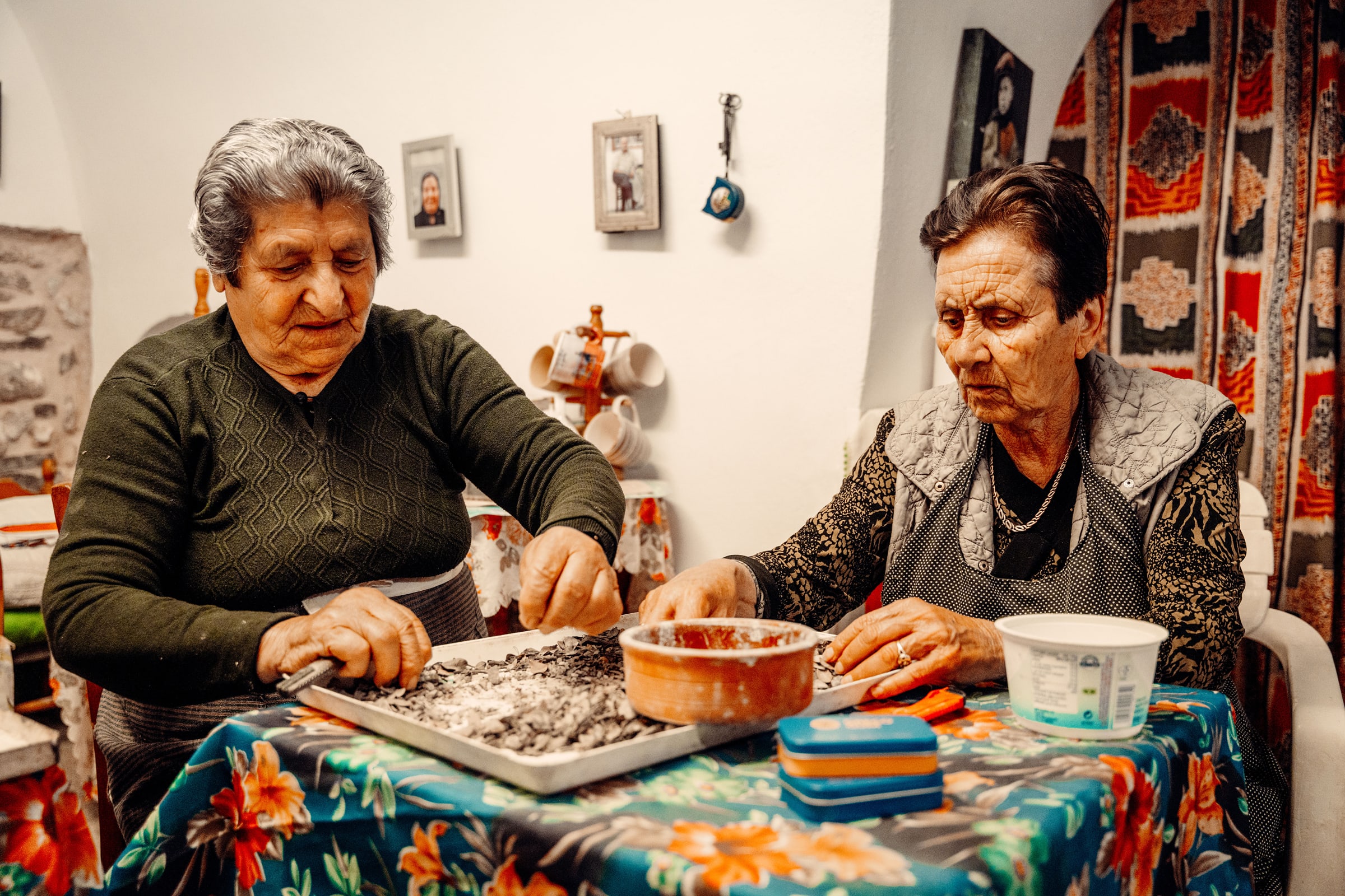 Two elderly women clean and sort mastic gum, with two tins of Greco Gum stacked in front of them