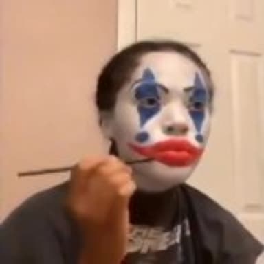 Clown Makeup Slander meme template: A person is applying clown makeup, with one eye already painted blue and white, and the lips painted red.