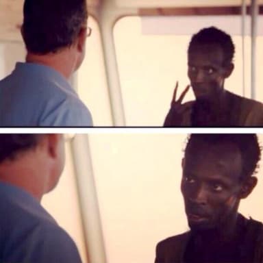 Captain Phillips I'm The Captain Now meme template: Two panels. Top panel: A man in a blue shirt turns away from the camera, looking out a window. A man in a dark shirt is visible through the window, making a peace sign. Bottom panel: Same scene, but the man in the dark shirt now looks intently at the camera.