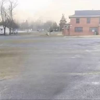 Blank Sybau meme template: A blurry, nondescript outdoor scene, possibly a parking lot or empty field, with some buildings in the background and trees on the left. The overall image is hazy and indistinct.