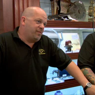 BEST I CAN DO IS meme template: Two men in black polo shirts standing behind a counter in what appears to be a pawn shop. The man on the left is bald and gesturing with his left hand, while the man on the right looks down with a neutral expression.