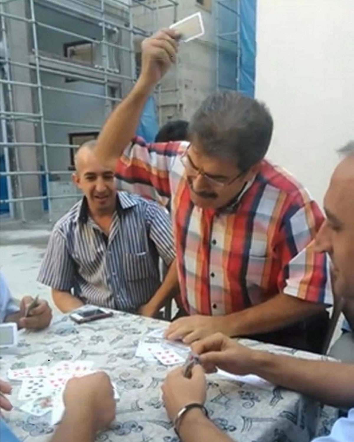 Angry Turkish Man Playing Cards meme template: A group of men are playing cards at a table, with one man in a red and orange plaid shirt holding up a card with a triumphant or frustrated expression.