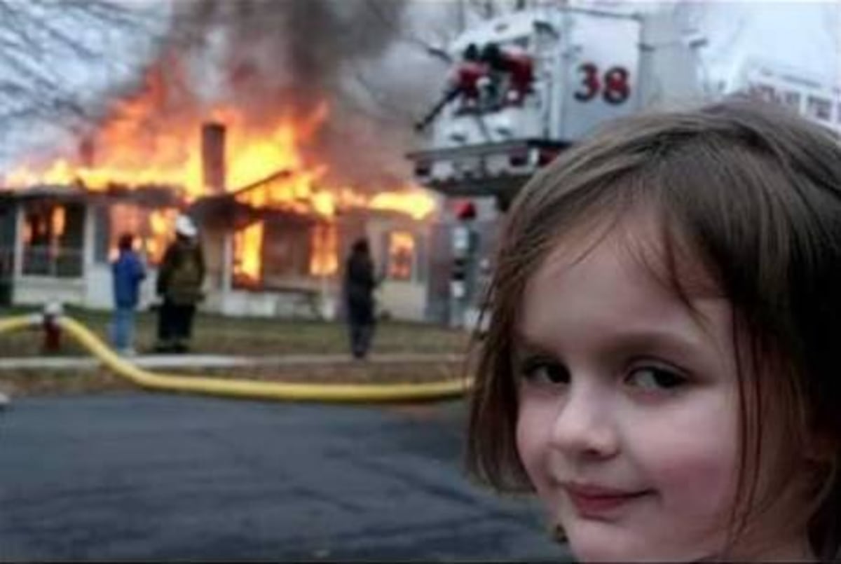 Burning House Girl meme template: A young girl with a mischievous smile looks towards the camera, while in the background a house is engulfed in flames and a fire truck is present.