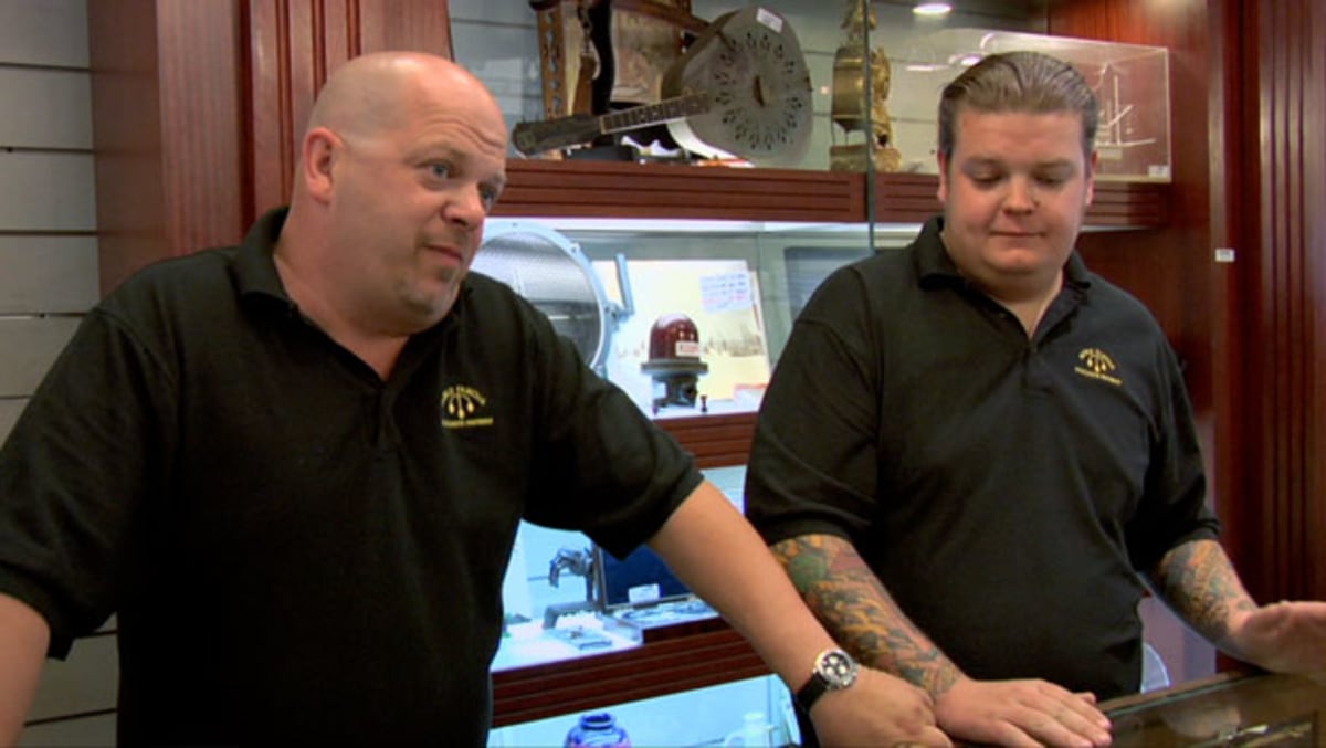 BEST I CAN DO IS meme template: Two men in black polo shirts standing behind a counter in what appears to be a pawn shop. The man on the left is bald and gesturing with his left hand, while the man on the right looks down with a neutral expression.