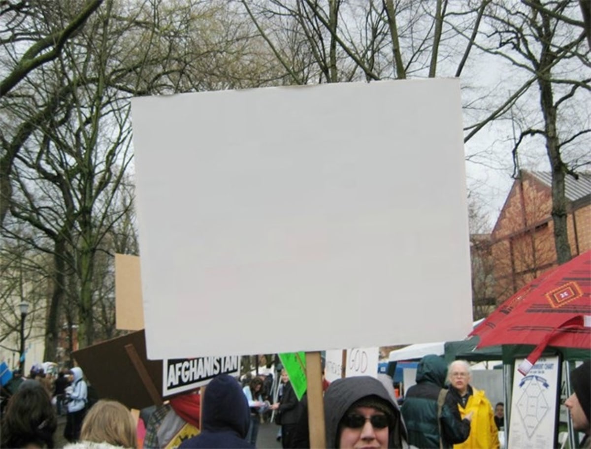 Blank Protest Sign meme template: A blank white protest sign is held up in front of a crowd. Other signs and tents are visible in the background.