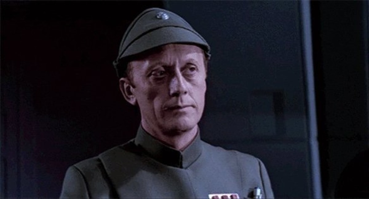 An Older Code Sir, But It Checks Out meme template: A man in a grey uniform and cap, with a stern expression, looking slightly to the side.