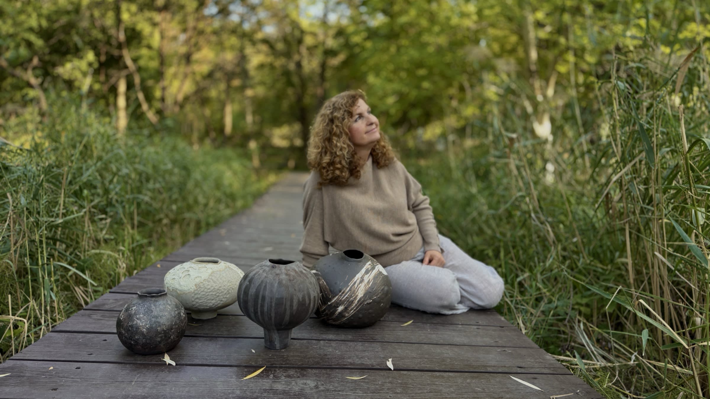 Helena sedí na drevenom chodníku v tráve pri svojich ručne modelovaných vázach