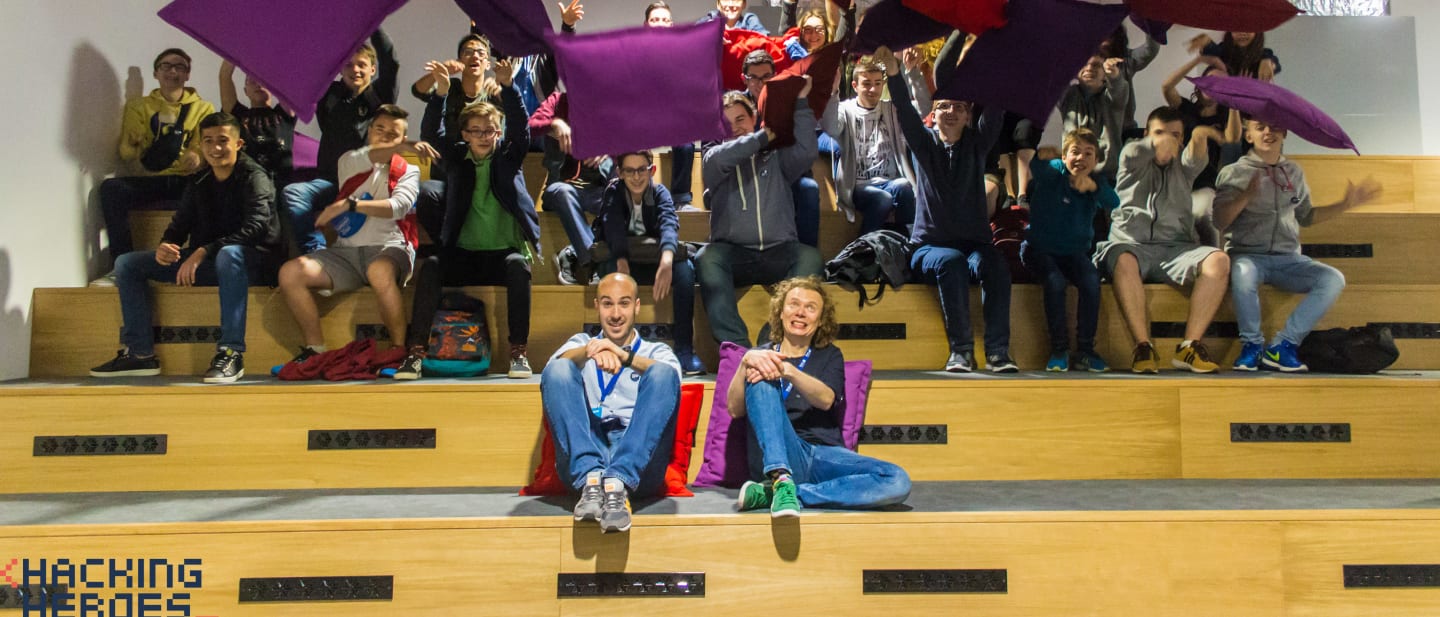 The whole group on tiered wooden seating, throwing cushions in the air, with two organisers sitting on the floor in front