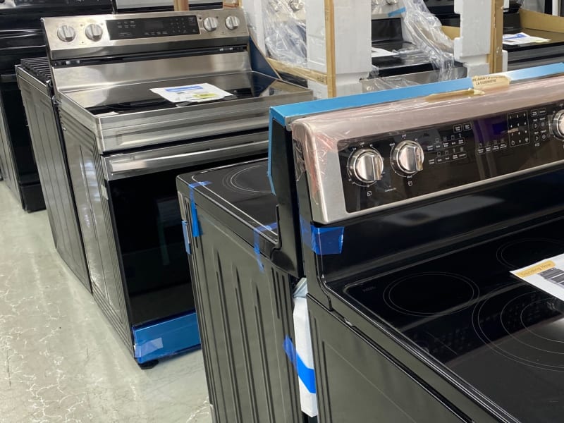 An aisle of new electric and gas ranges, some still in packing foam, with refrigerators behind them at Barn Appliances in Leoma