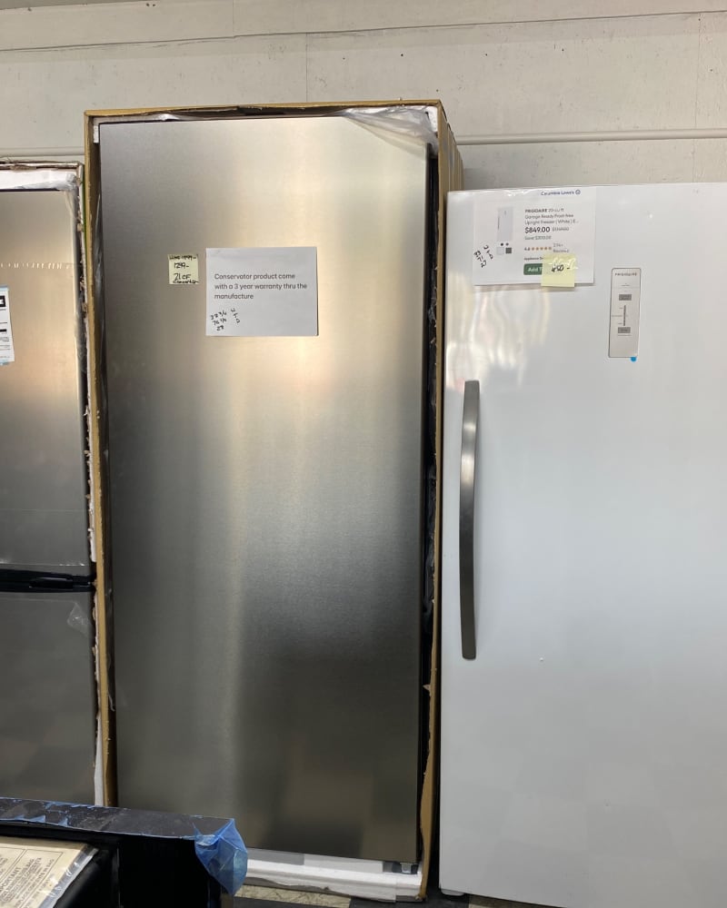 A stainless upright freezer and a white Frigidaire garage-ready upright freezer standing in their cartons at Barn Appliances