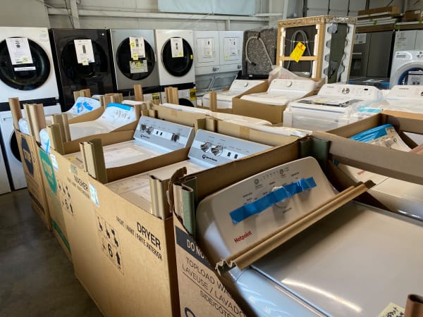 Speed Queen and Hotpoint washers in their shipping boxes in front of stacked LG laundry centers