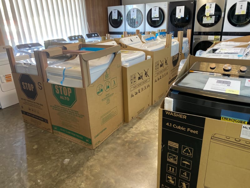 Top-load washers and dryers in their cartons beside stacked LG laundry centers in the wood-paneled laundry room at Barn Appliances