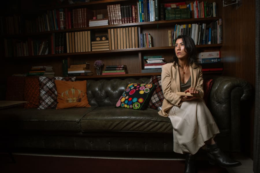 Mulher de blazer bege sentada em sofá de couro, em sala com estantes de livros ao fundo