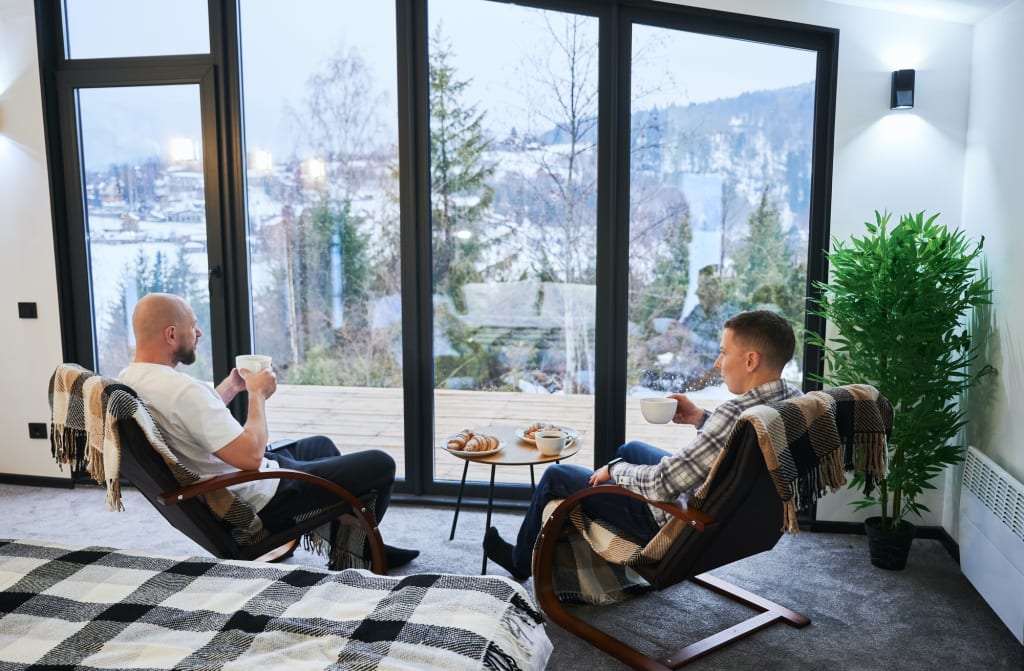 Two young men drinking tea, enjoying view of winter mountain landscape through panoramic windows.