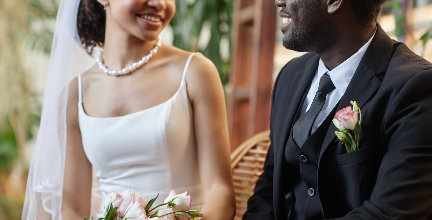 A bride and groom smile at each other on their wedding day, holding flowers and dressed in formal attire. The joyful moment contrasts the unseen issue of an unrecorded marriage license; a situation w