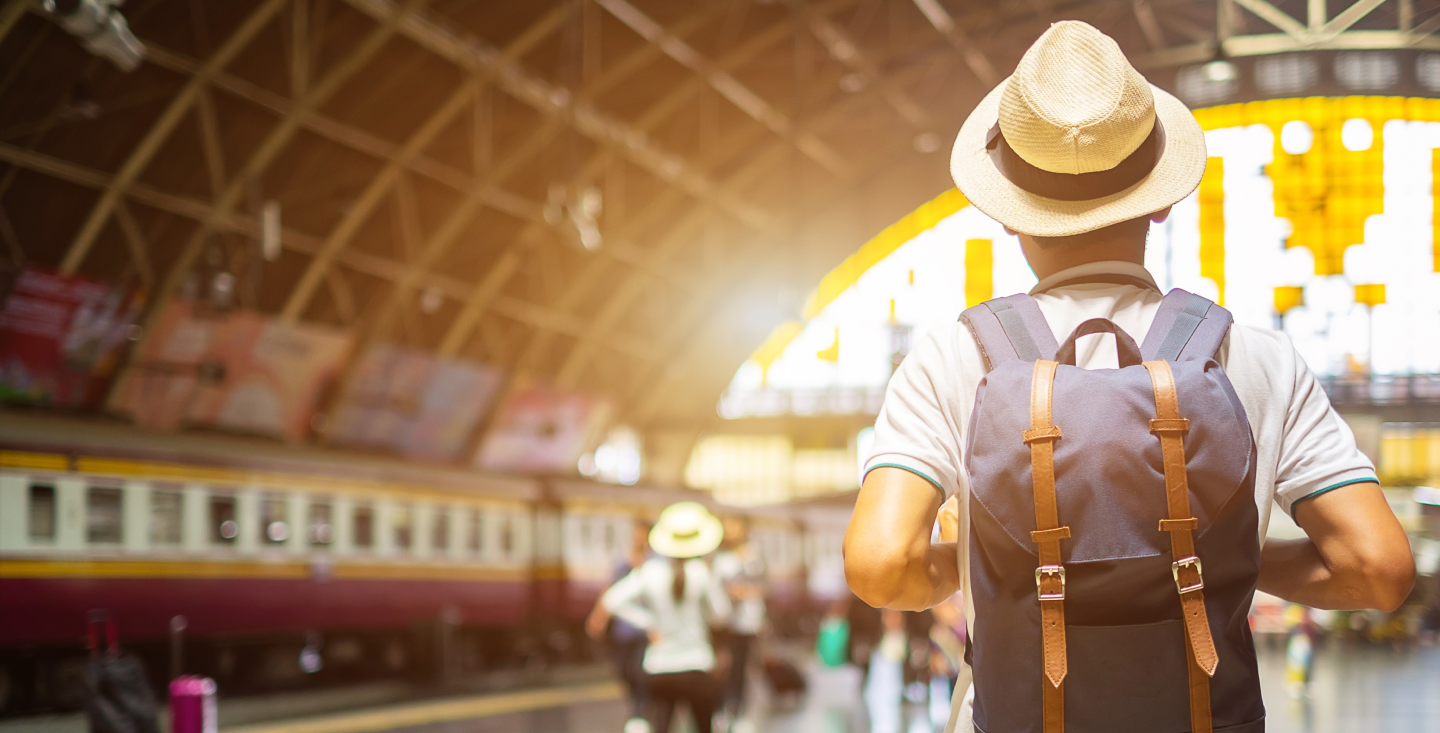 Modern traveler in a straw hat and backpack waiting at a bright, bustling train station, ready for the next adventure.