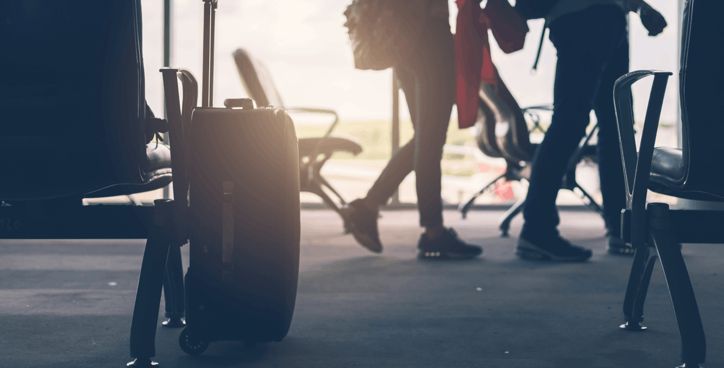 Silhouettes of travelers walking through an airport terminal with carry-on luggage and backpacks, with sunlight streaming through large windows
