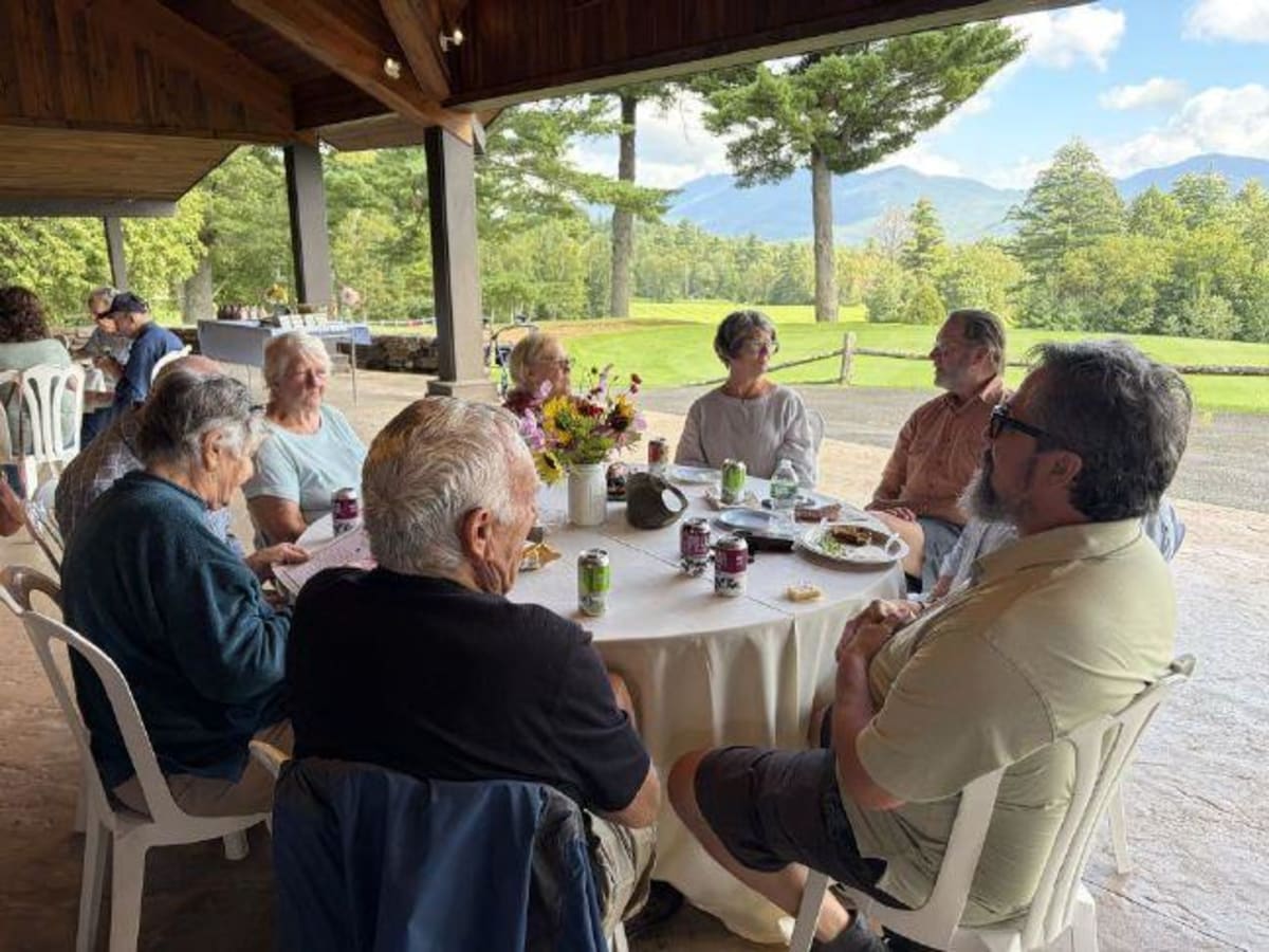 ** Volunteers and elderly friends share lunch and conversation at outdoor community picnic