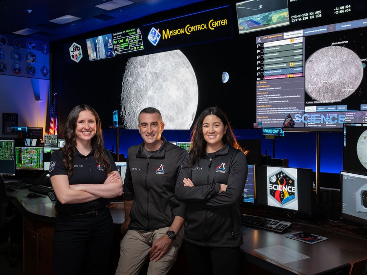Three NASA science officers in business attire standing beside Mission Control console with multiple monitors