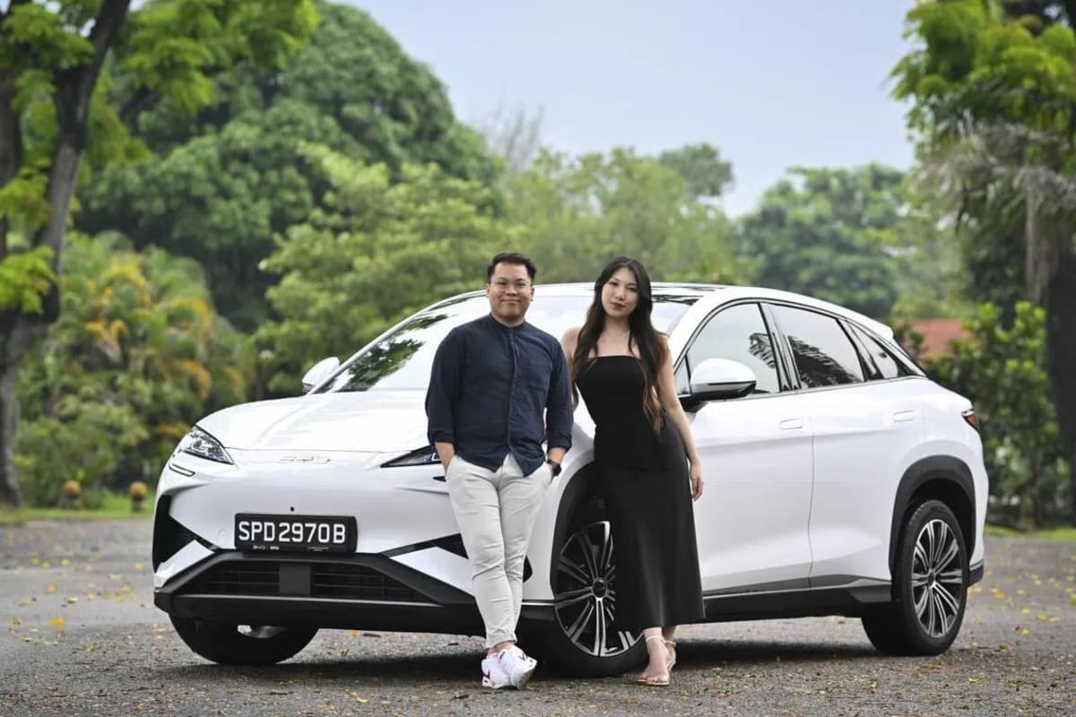Young couple standing beside their white electric vehicle in Singapore, smiling at camera