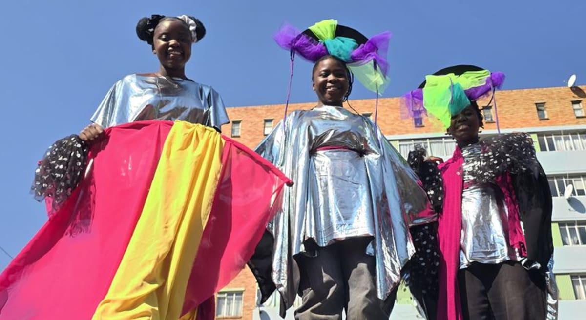 Three young girls in vibrant handmade bird costumes smile at Hillbrow Carnival in Johannesburg