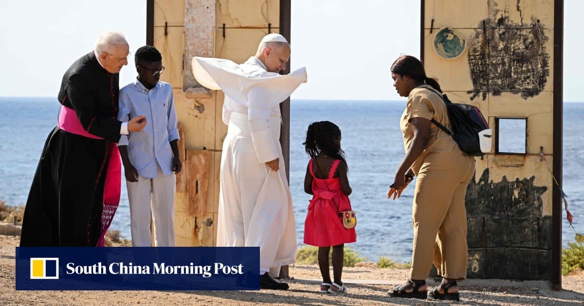 Pope Leo standing on rocky Lampedusa shoreline holding hands with migrant children and their mother