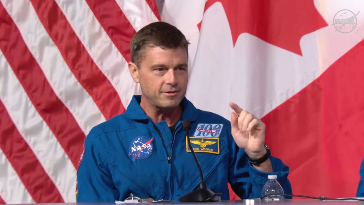 NASA astronaut Reid Wiseman in blue flight suit speaking to reporters at Johnson Space Center