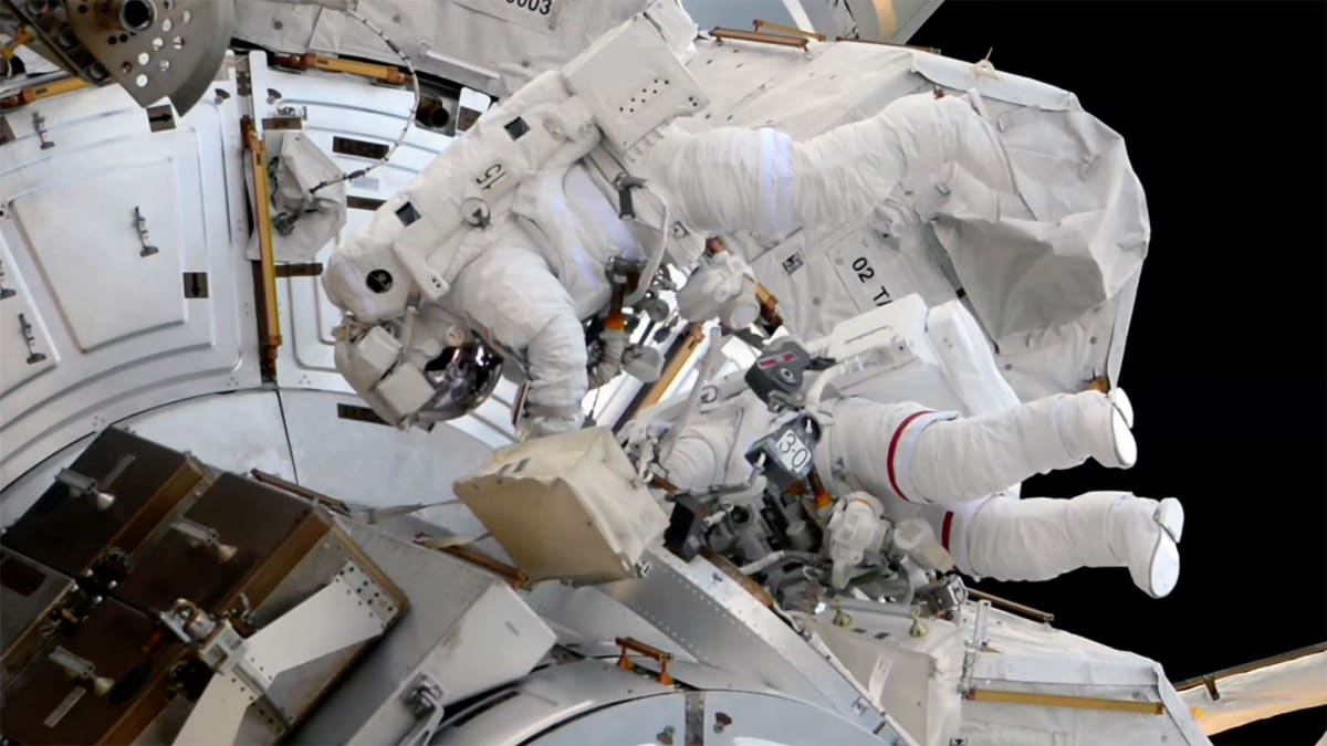 Two astronauts in white spacesuits work outside International Space Station against black space backdrop