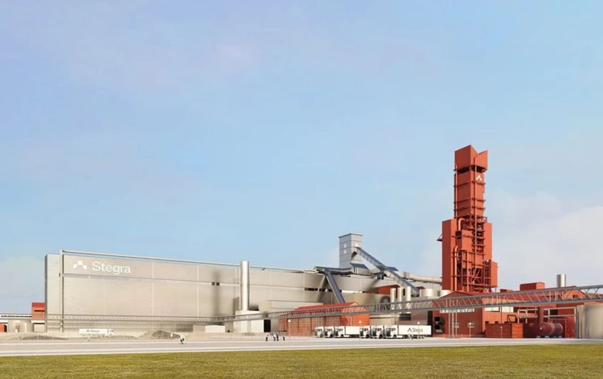 ** Modern industrial steel manufacturing facility with wind turbines visible in background representing green energy production
