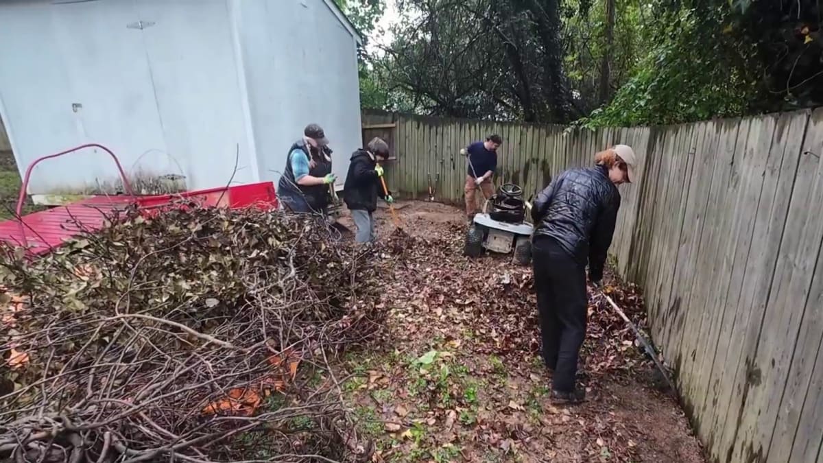 Volunteers rake leaves and trim overgrown bushes in elderly couple's yard on rainy day