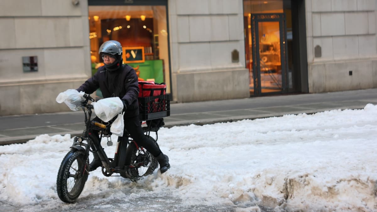 Modern glass and metal shelter on Manhattan sidewalk with delivery workers in bike helmets gathering nearby