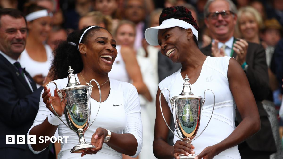 Venus and Serena Williams holding Wimbledon doubles trophy together on tennis court