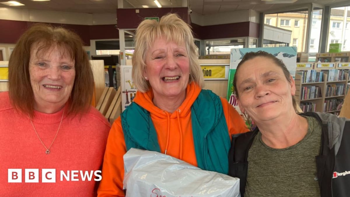 Three smiling women stand together surrounded by organized school uniforms in charity warehouse