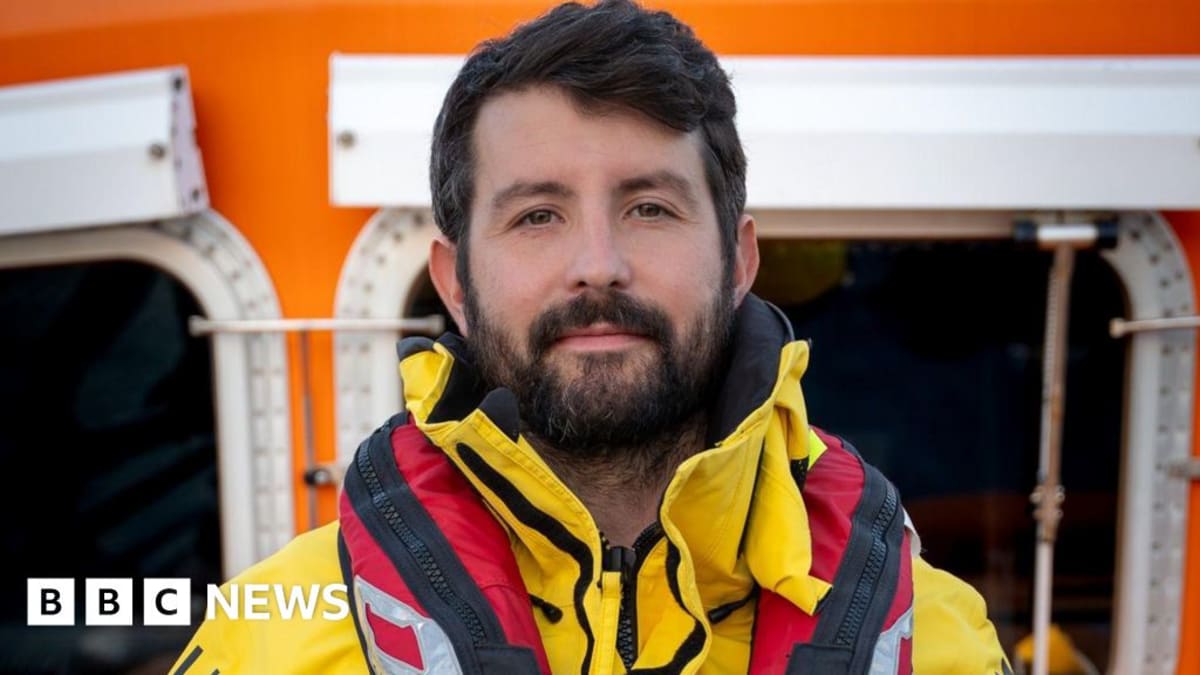 RNLI volunteer Will Roberts in orange rescue gear smiling at camera after maritime rescue mission