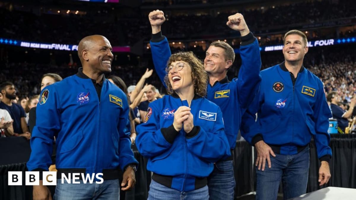 Four Artemis II astronauts in blue flight suits celebrating with crowd at Houston basketball game