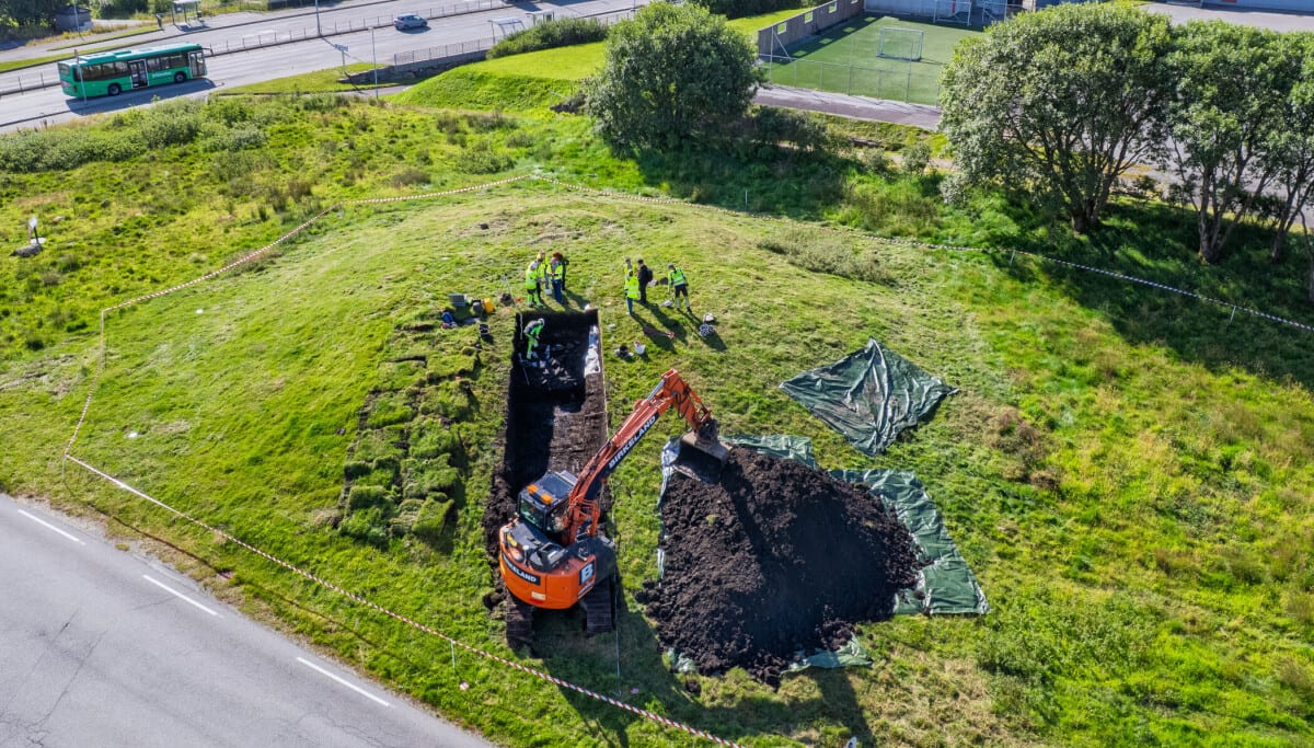 ** Aerial view of archaeological excavation trench cut into large green burial mound at Grønhaug, Norway