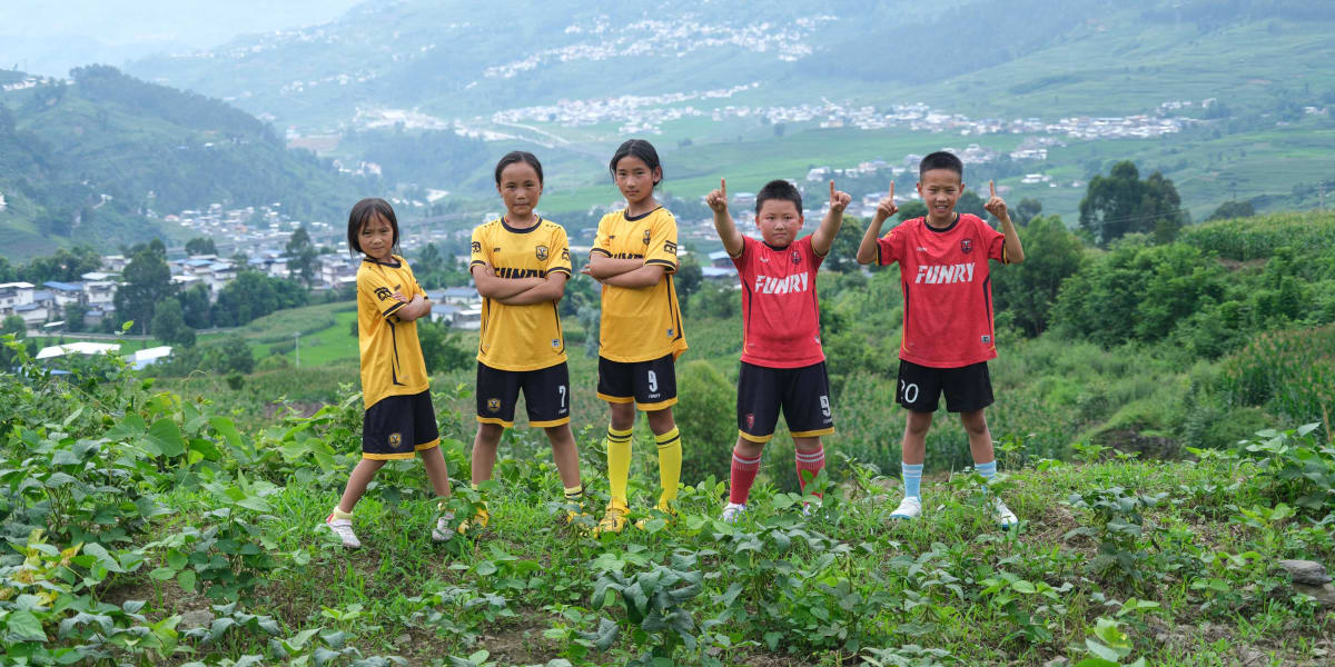 Young children in soccer uniforms walking through mountain rice paddies toward training pitch