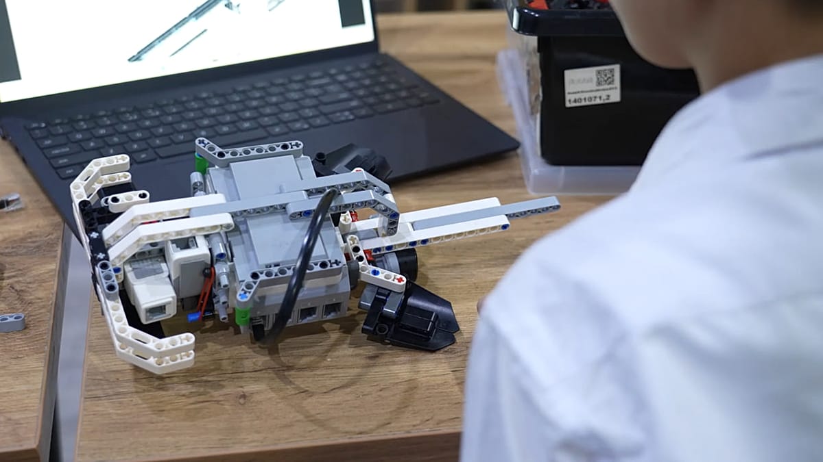 Young Uzbek student programming educational robot at desk with laptop in classroom
