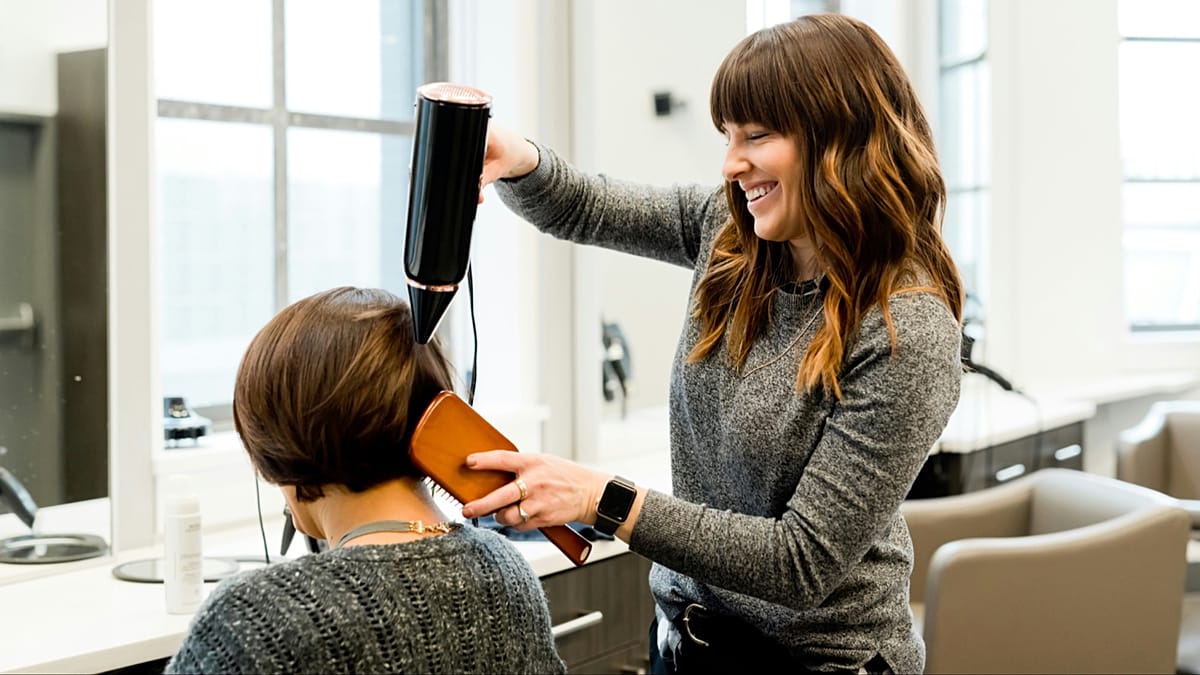Hairdresser styling client's hair in modern salon with sustainability message visible on mirror